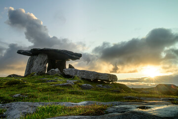 The dolmen portal tomb of Poulnabrone with the golden sun rising in a grey, cloudy sky over the Burren; County Clare, Ireland
