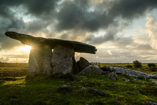 The dolmen portal tomb of Poulnabrone with the golden sun rising in a grey, cloudy sky on the Burren; County Clare, Ireland