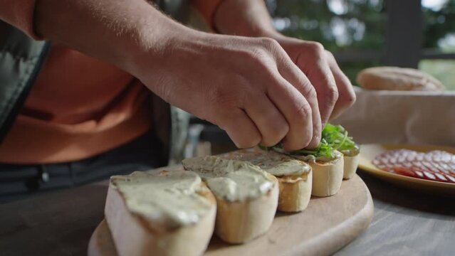 Close up shot of hands of male chef adding arugula leaves to bruschettas while cooking appetizers on outdoor terrace