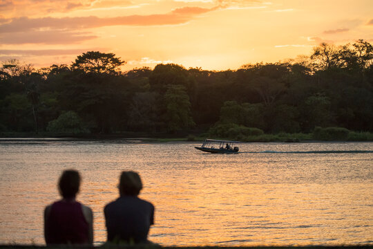 View from behind of a silhouette of tourists sitting on the shore watching a golden sunset with a boat traveling the waterways that surround Tortuguero; Limon Province, Costa Rica