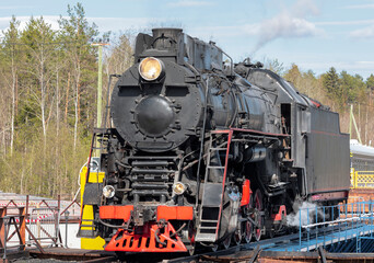 Black steam locomotive with a burning spotlight on a turning circle against the backdrop of a green forest