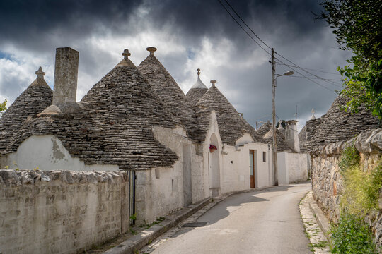 Street scene with a row of traditional Apulian round stone Trulli houses with a stormy sky on a sunny day in the town of Alberobello; Alberobello, Puglia, Italy