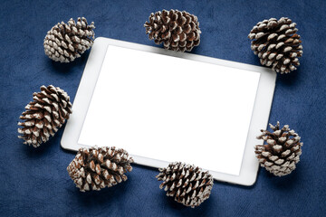 mockup of digital tablet with a blank isolated screen (clipping path included), flat lay with decorative, frosty pine cones on blue textured paper, winter holidays theme