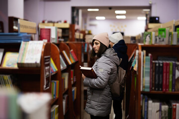 Mother with son in jacket and backpack reaching a book from bookshelf at the library.
