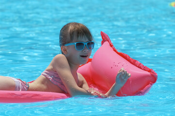Young joyful child girl having fun swimming on inflatable air mattress in swimming pool with blue...