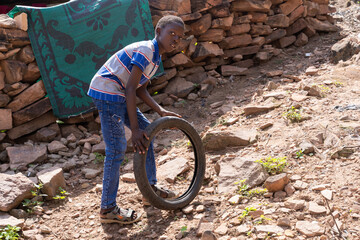 Handsome African youngster playing with his old bicycle wheel on the steep rough road in front of...