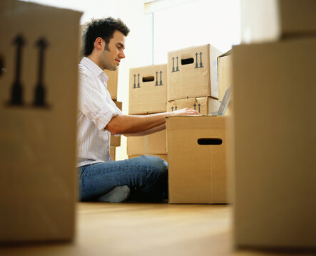 A man sits working on a laptop propped on a moving box, surrounded by packed boxes in a home environment; Germany