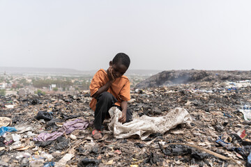 Miserable hungry African slum child looking for edible and recyclable items in a huge urban garbage...