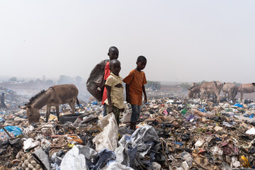 Three African street kids standing in a steaming dump looking for recyclable items to sell - obrazy, fototapety, plakaty