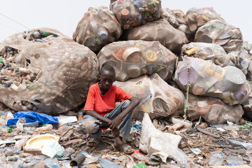 Young African boy in a red t-shirt kneeling thoughtfully in front of a huge pile of recycled...