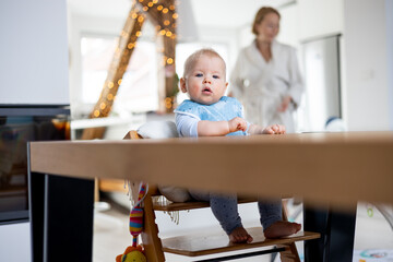 Happy infant sitting at dining table and playing with his toy in traditional scandinavian designer wooden high chair in modern bright atic home superwised by his mother.