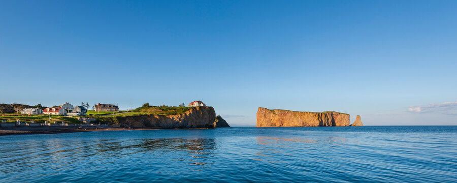 Panoramic view of Perce Rock and house on cliff edge with bright blue sky, Gaspe Peninsula; Quebec, Canada