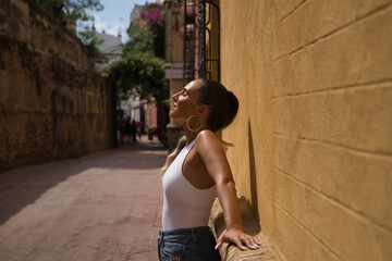 Young and beautiful woman, with ponytail, wearing a white top and jeans, with her eyes closed, leaning against a wall, relaxed, receiving the sun's rays. Concept beauty, sun, relax, peace, tranquility