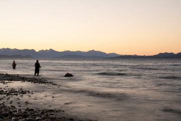 Fototapeta premium Night shot. View of the water blurred effect and blurred fishermen dark silhouette, fishing at nightfall from the coast. The rocky shore and mountains in the horizon with a beautiful dusk light.