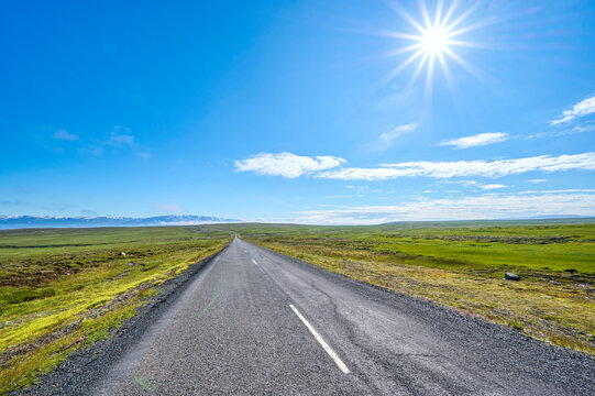 Vast empty road in summer with sunburst in bright blue sky overhead; Austurland, Northern region, Iceland