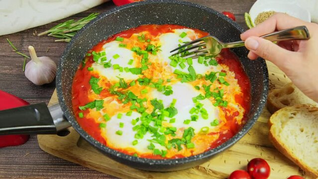Person eating shakshuka, tomato eggs served in pan on wooden table. hands cutting poached eggs in tomatoes with fork, spicy shakshouka, traditional Israeli breakfast, slow motion video, 4k fottage