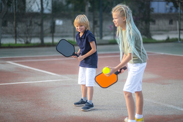 Laughing boy and girl playing pickleball game, hitting pickleball yellow ball with paddle, outdoor...