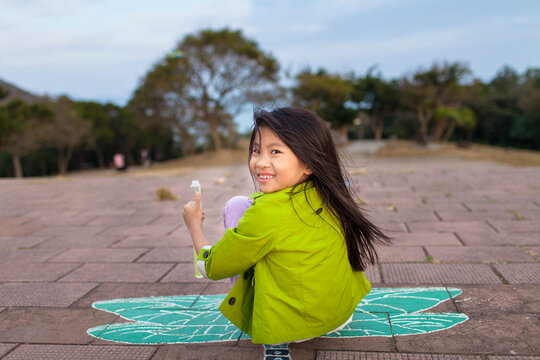 Young girl sits on the ground on a painted butterfly, looking back at the camera and giving a thumbs up; Hong Kong, China