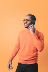 The portrait of a handsome Colombian man making a call with his cell phone, the man is wearing a shirt in the same color as the background. orange background.