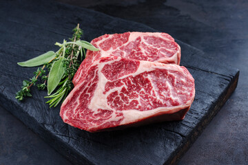 Two raw dry aged wagyu rib-eye beef steaks with herbs offered as close-up on a rustic charred...