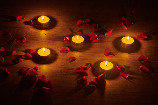 Illuminated tealight candles and red rose petals scattered on a surface create a romantic setting; Studio
