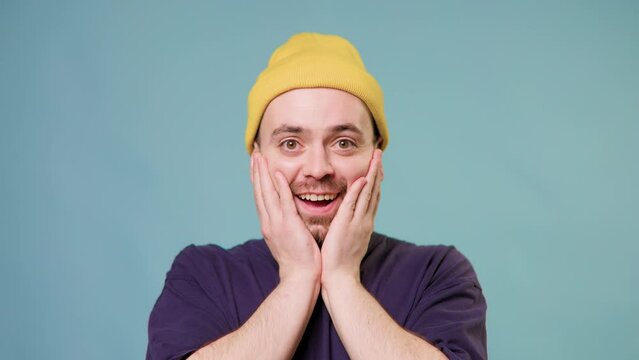 European Amazed hipster guy pleasantly surprised to camera over blue background Wow, unbelievable, opening mouth in amazement, shocked man put hands on face and very surprised and looking at camera