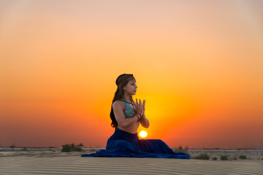 Woman sits on the desert sands against a glowing golden sunset in the yoga prayer pose; Abu Dhabi, United Arab Emirates