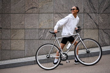 Joyful african american woman in sunglasses walking near bicycle on urban street 