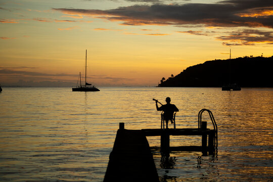 Man sitting on a dock playing the banjo at sunset with anchored sailboats off the shoreline; Tahaa, French Polynesia