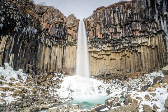 Cascata Svartifoss, torrente d'acqua che cade da un salto di rocce basaltiche. Sassi sul letto del torrente, ghiaccio alla base della cascata, stalattiti di ghiaccio