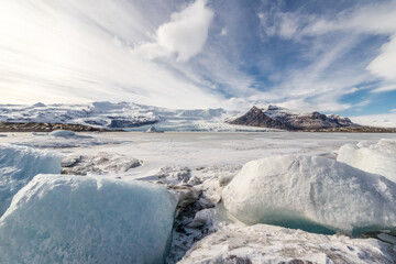 laguna del Fjallsárlón illuminata dal sole dietro delle nuvole lattiginose, la lingua del ghiacciaio termina direttamente nella laguna scendendo dal versante della montagna, in primo piano due iceberg