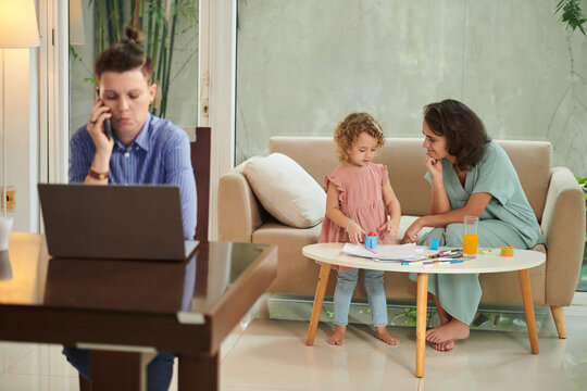 Smiling woman playing with little daughter when wife working on laptop at home