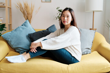 Side view of young female in casual clothes sitting on sofa and looking at camera while resting in cozy living room