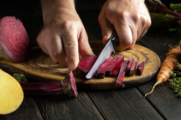 Close-up of a chef hands with a knife cut red table beets on a kitchen cutting board before...