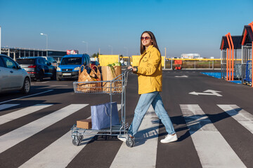 Fototapeta na wymiar Woman in yellow jacket goes crosswalk from a shopping mall with shopping cart with a groceries