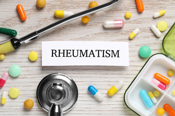 Card with word Rheumatism, stethoscope and pills on white wooden table, flat lay