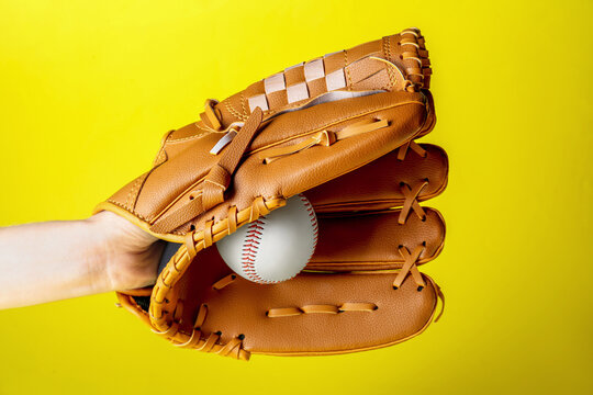 Baseball player holding ball with catcher's mitt on yellow background, closeup. Sports game