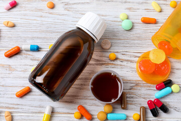 Bottle of syrup, measuring cup, lozenges and pills on white wooden table, flat lay. Cold medicine