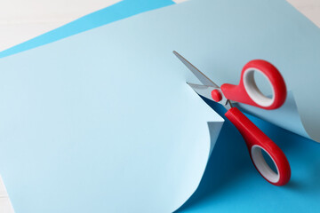 Red scissors cutting light blue paper on white background, closeup