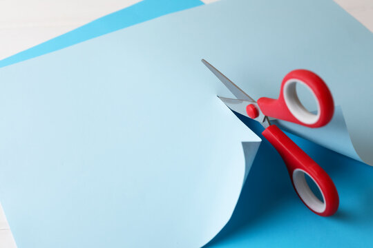 Red scissors cutting light blue paper on white background, closeup