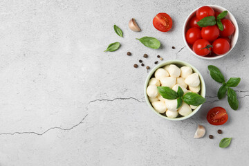 Delicious mozzarella balls in bowl, tomatoes and basil leaves on light gray textured table, flat lay. Space for text