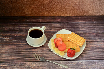 A cup of hot coffee and a plate of fresh waffles with strawberries and bananas on a wooden table.