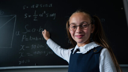 Portrait of a caucasian girl in glasses in the classroom. The schoolgirl writes the formula with chalk on the blackboard and looks into the camera.