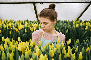 Portrait of young beautiful woman standing in hothouse and enjoying smell of yellow tulips with her eyes closed  