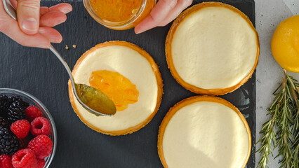 Topping cheesecake with orange marmalade. Mini cheesecakes, and ingredients for the dessert topping close-up on kitchen table, woman decorating cheesecake
