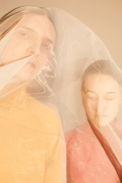 Young man and woman in unisex sweaters keeping eyes closed and looking at camera while being covered with translucent material