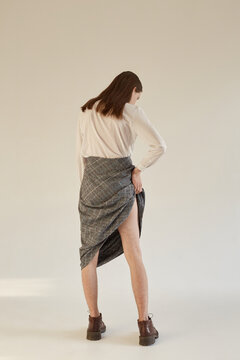Back view of slim androgynous man in female outfit pulling up long skirt and showing leg while standing against gray background