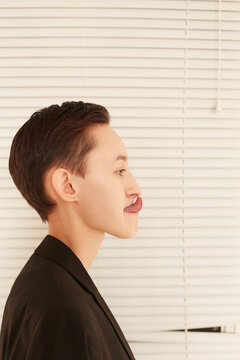 Side view of pretty androgynous female in male suit touching nose with tongue while standing near window covered with blindsSide view of pretty androgynous female in male suit touching nose with tongu
