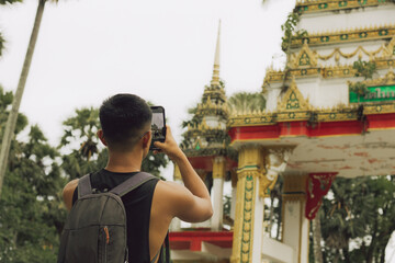 Back of young man taking photos with smart phone of Buddhist temple in Phuket. Male tourist visiting religious building in Thailand. Backpacker travel in South East Asia concept