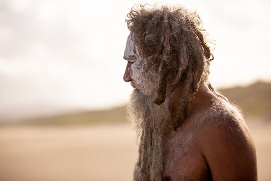 Aboriginal middle aged man with clay face paint standing on a beach looking into the distance
